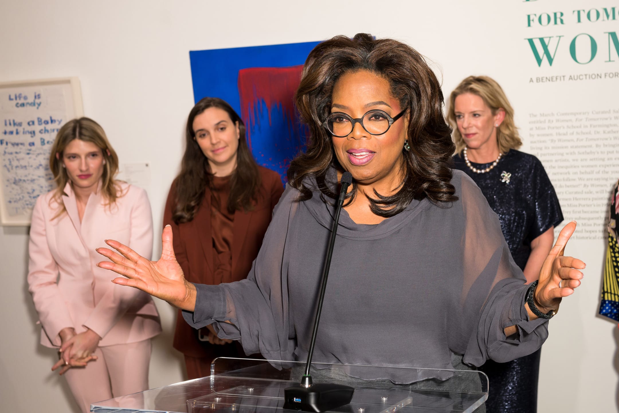 A woman speaking at a podium with an audience in the background at an art event.