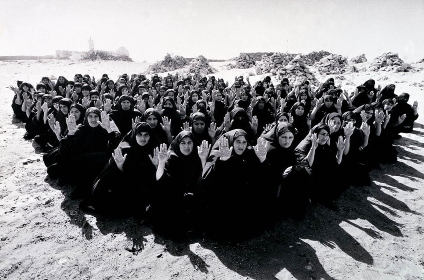 A group of women in black clothing sit outdoors on sandy ground, facing the camera with hands raised in unison.