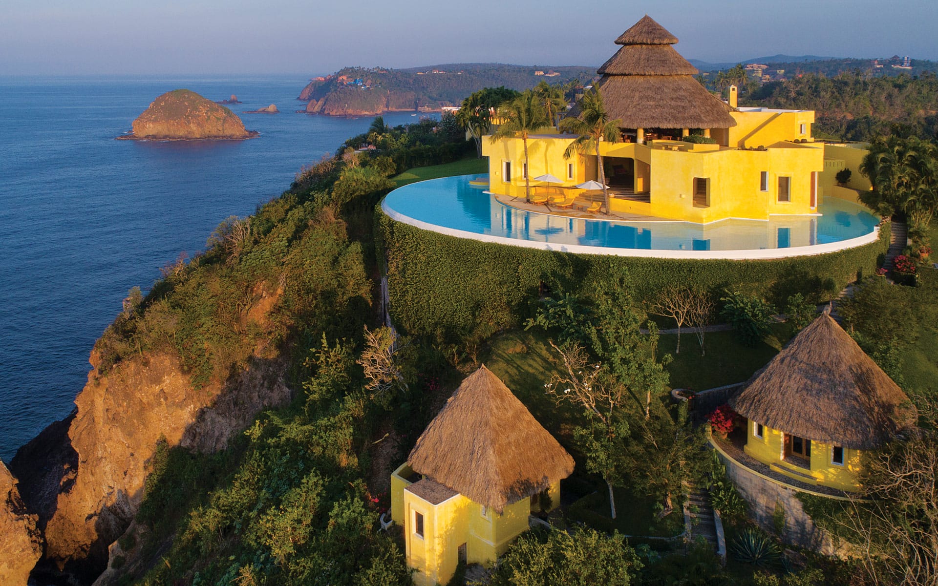 Luxurious cliffside villa with thatched roof and infinity pool overlooking the ocean at sunset.