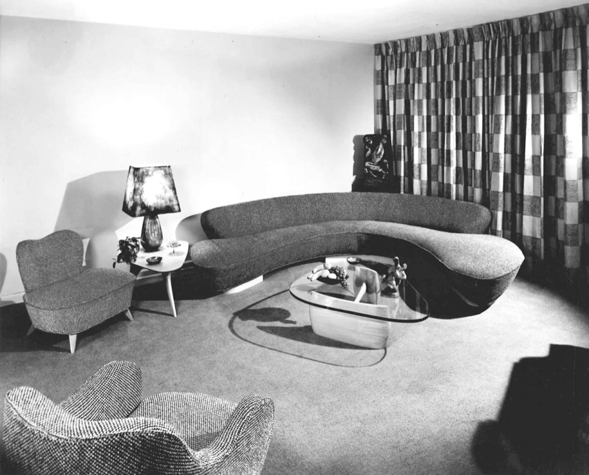 Mid-century modern living room with curved sofa, patterned curtains, armchairs, a coffee table, and decorative lamp.