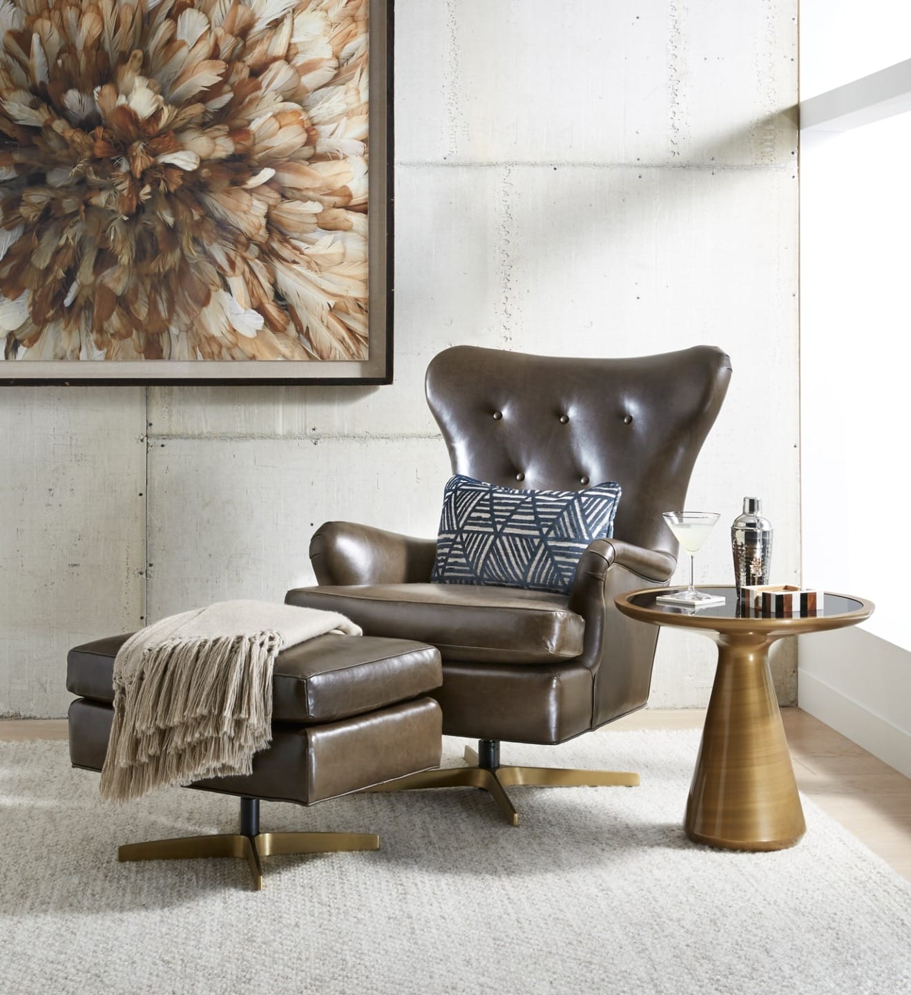 Brown leather armchair with ottoman next to a round table, abstract artwork, and martini glass on a light carpeted floor.