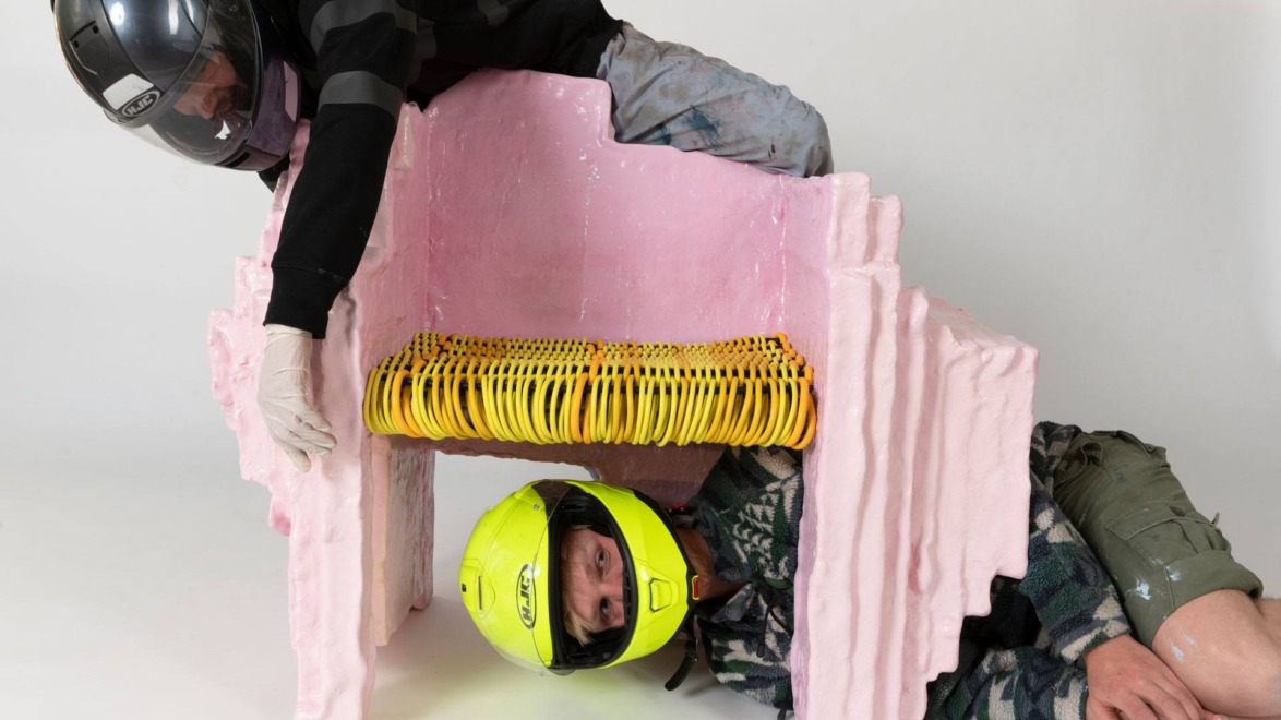 Two people in helmets interact with an abstract pink sculpture, one standing and the other peeking through an opening.