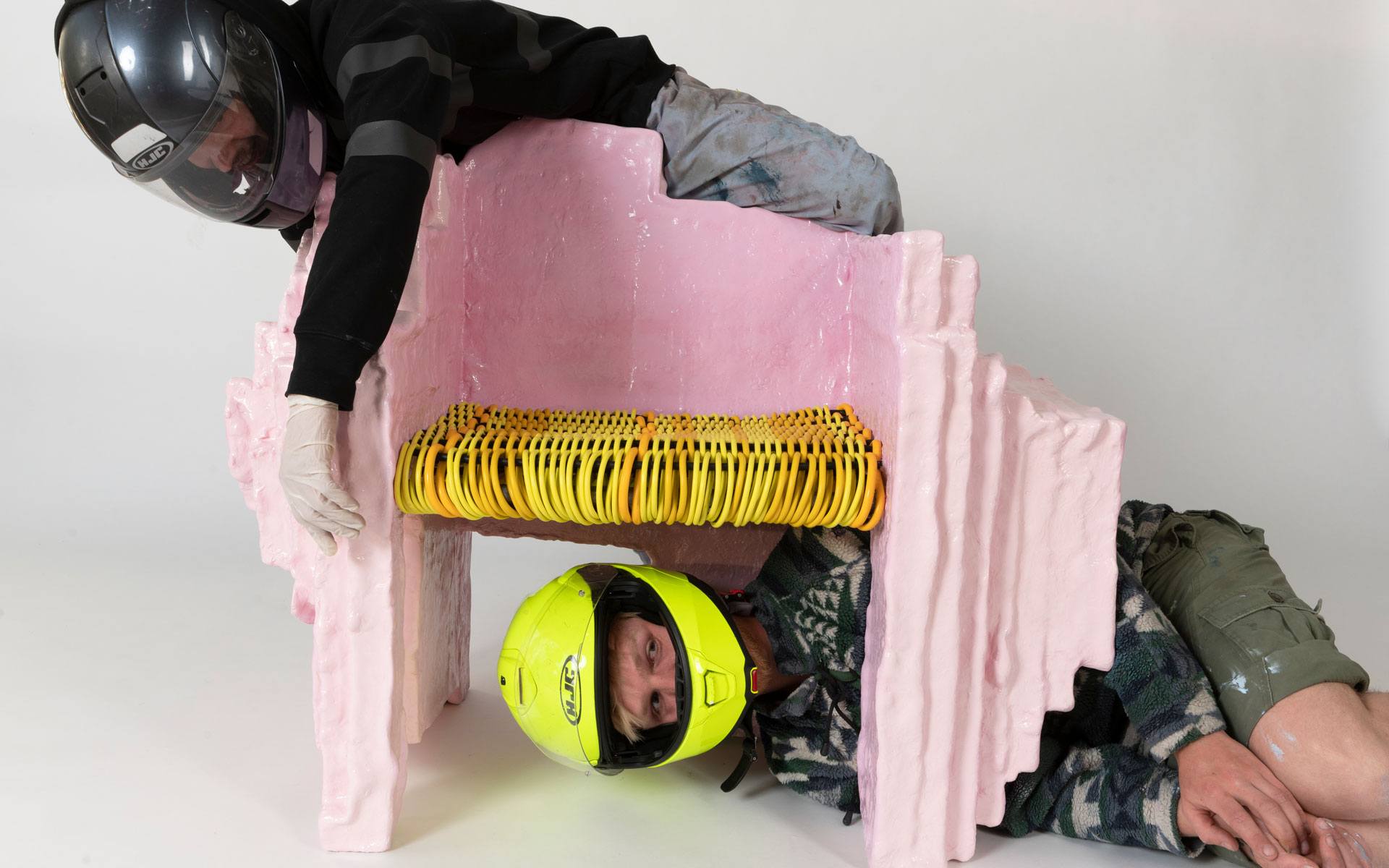 Two people in helmets interact with an abstract pink sculpture, one standing and the other peeking through an opening.