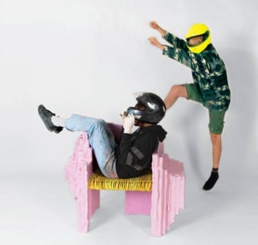 Two people with helmets, one sitting in a pink chair, the other jumping, wearing casual clothes, on a white background.