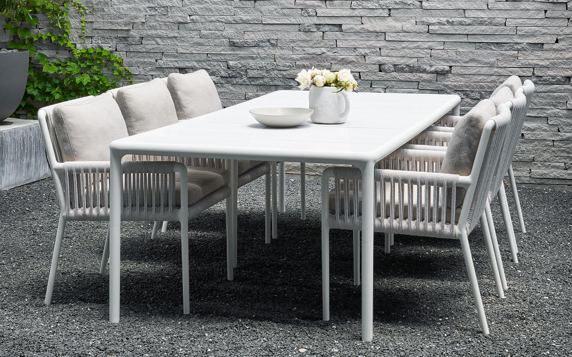 White outdoor dining set with a table and six chairs on a gravel patio, against a stone wall with some greenery.