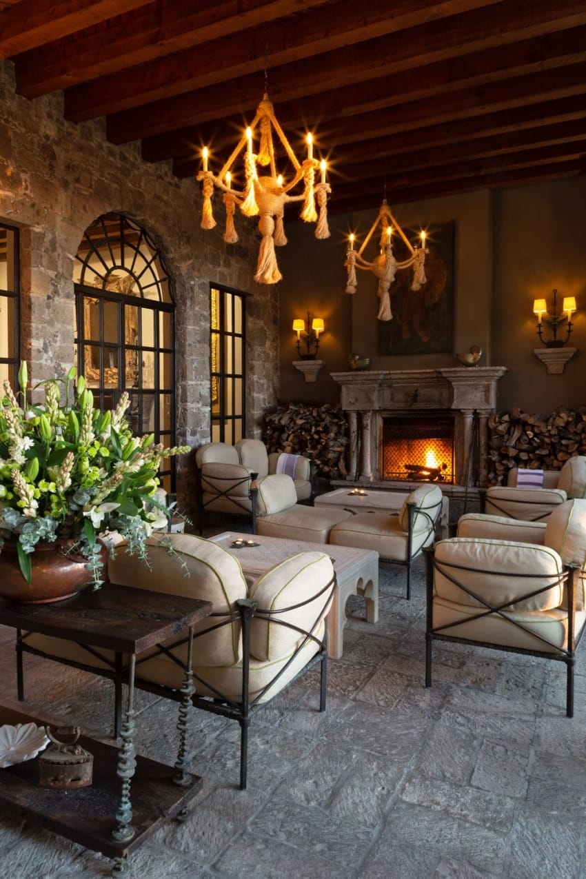 Rustic living room with stone walls, lit fireplace, wooden ceiling, and elegant chandeliers, surrounded by plush chairs and decor.