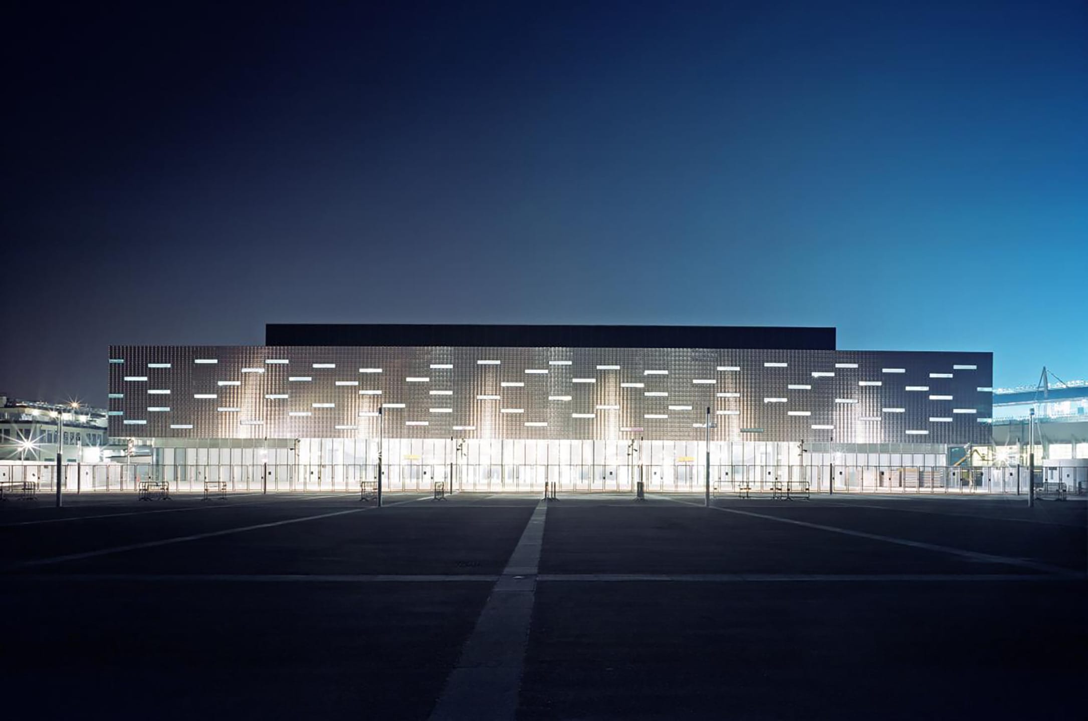 Modern building facade illuminated at night with a grid pattern and large windows reflecting on a dark surface