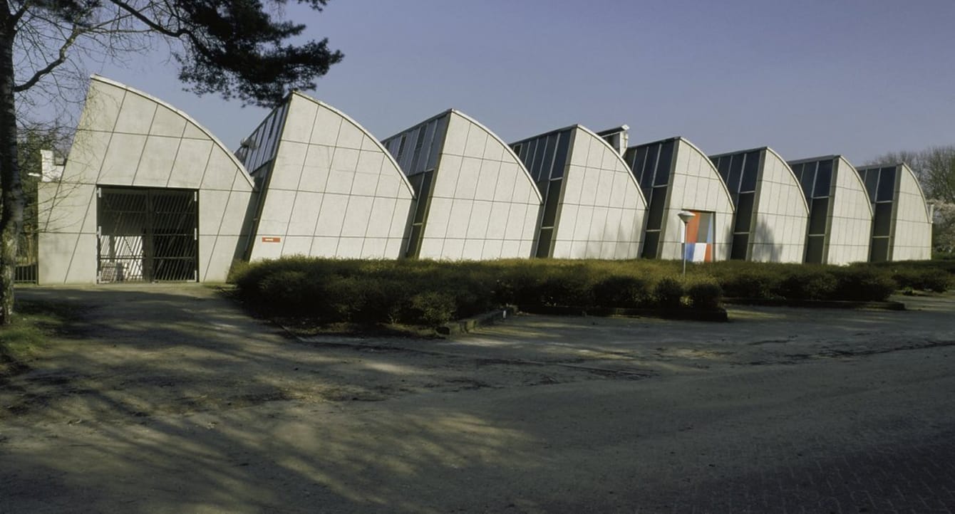 Modern white industrial building with distinctive curved roof sections and surrounding shrubs under a clear blue sky.
