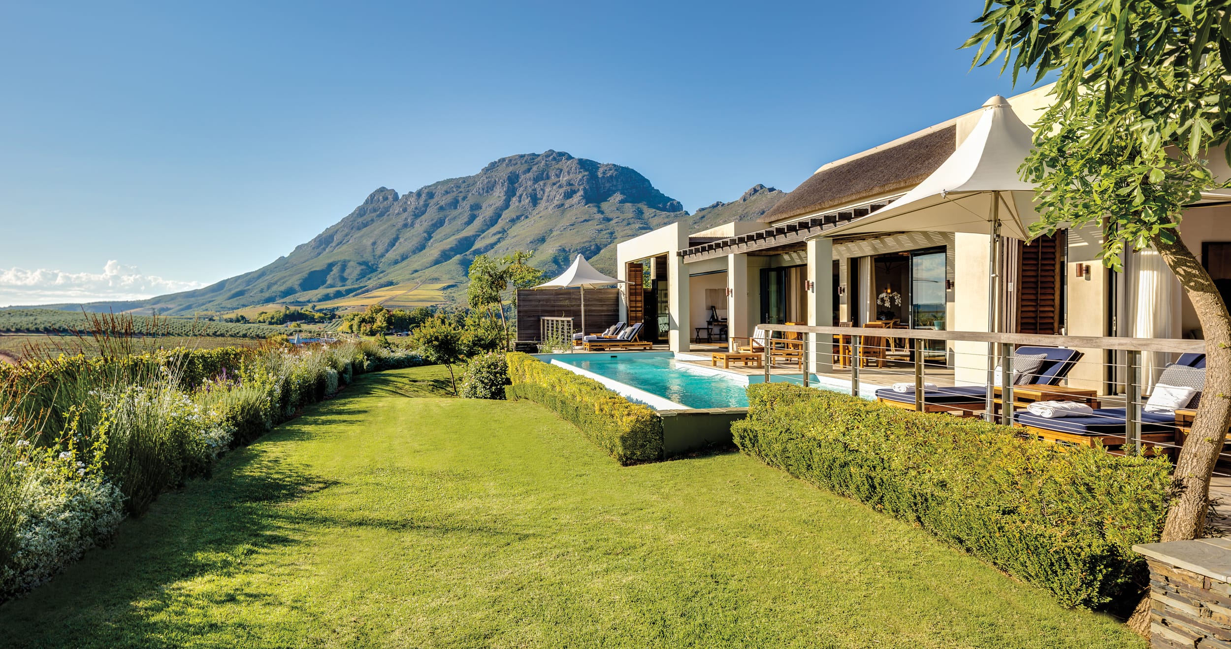 Luxury villa with a pool overlooking a lush garden and mountain view, under a clear blue sky.