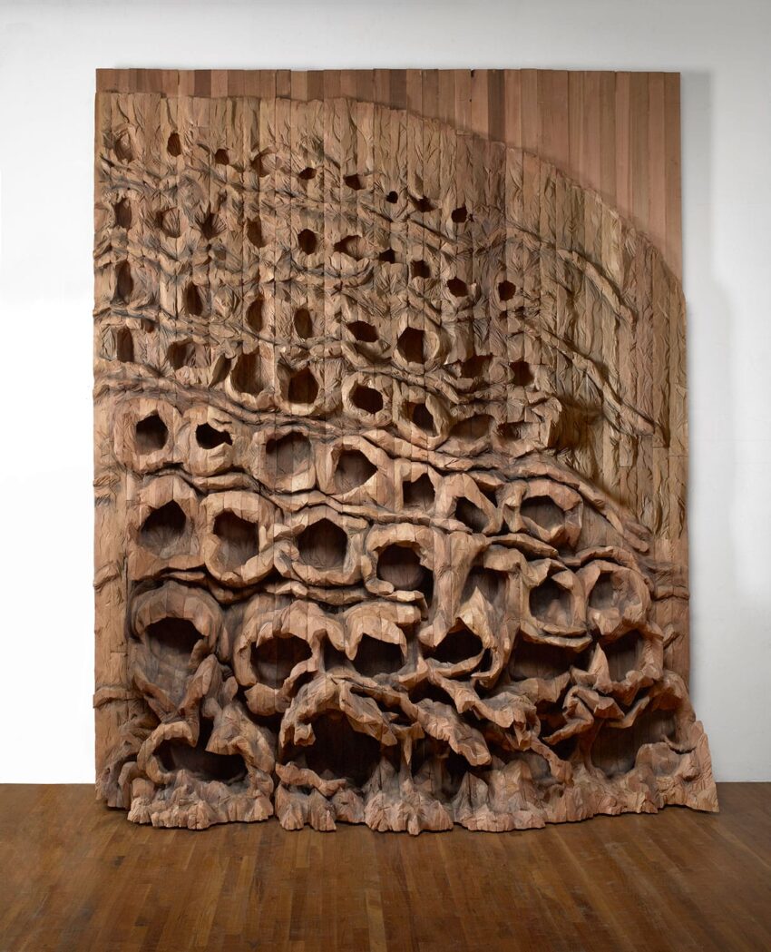 Large wooden sculpture with intricate, carved patterns resembling natural formations, displayed on a wooden floor.