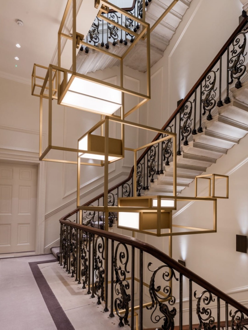 Modern geometric light fixture hanging in a stairwell with ornate black railings and a beige color scheme.