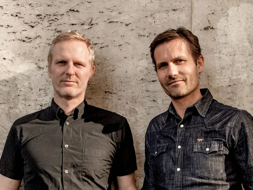Two men standing against a textured wall, both wearing dark shirts and looking at the camera.