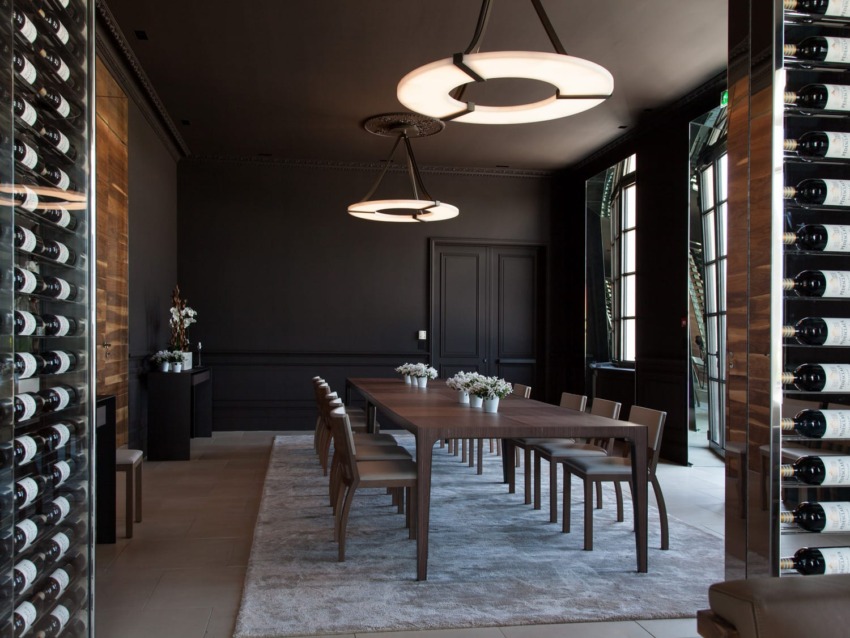 Modern dining room with a long wooden table, elegant chairs, contemporary lighting, and a wine display on the side wall.