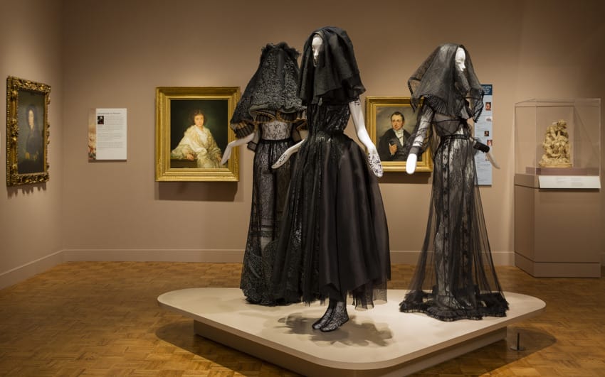 Three mannequins in black lace dresses and veils in an art gallery with paintings on the walls.