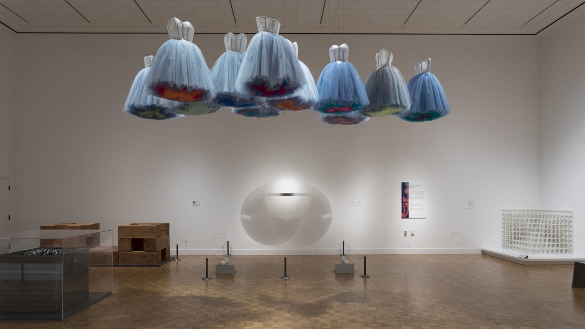 Suspended blue dresses art installation in modern gallery with wooden floor and minimal white walls.