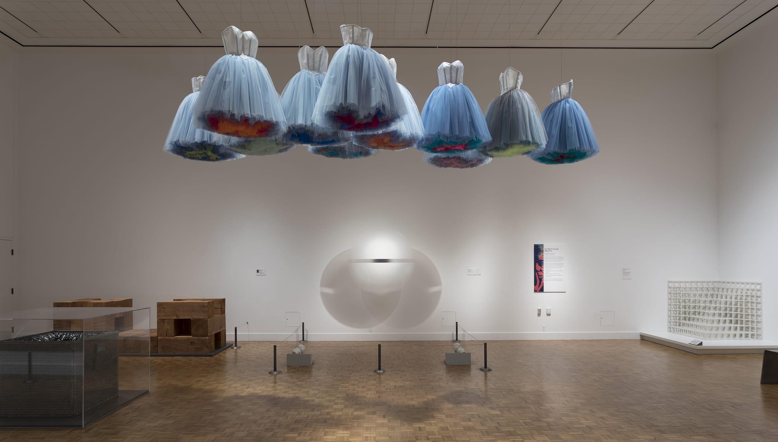 Suspended blue dresses art installation in modern gallery with wooden floor and minimal white walls.