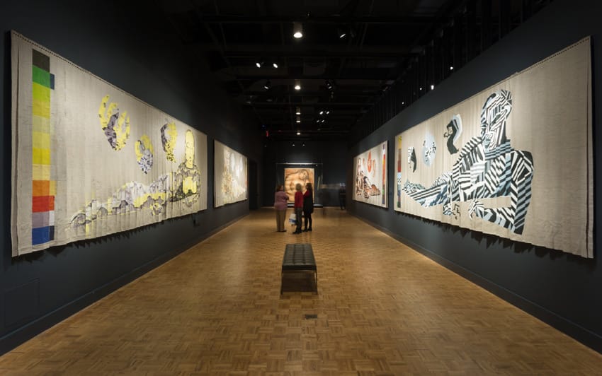 People observing large abstract paintings in a dimly lit gallery with wooden flooring.