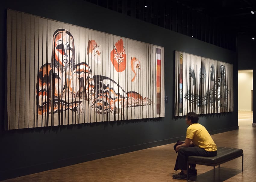 Man sitting and viewing large, abstract tapestries in a dimly lit art gallery.