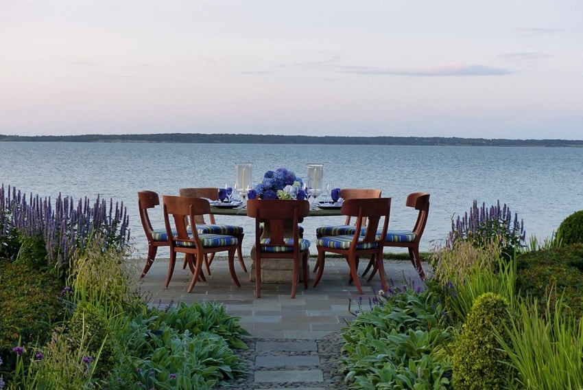 Outdoor dining table by a calm lake, surrounded by greenery and purple flowers, set for six with blue and white decorations.
