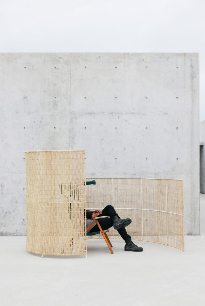 Person in black clothing sitting on a chair behind a woven screen against a concrete wall background.