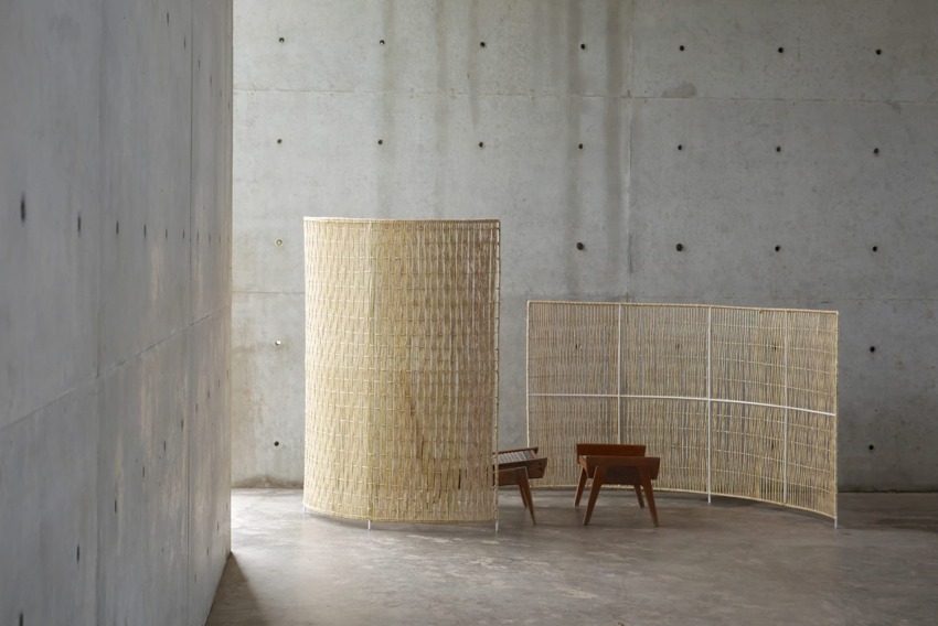 Minimalist room with two wooden chairs and curved woven room dividers against a gray concrete wall background.