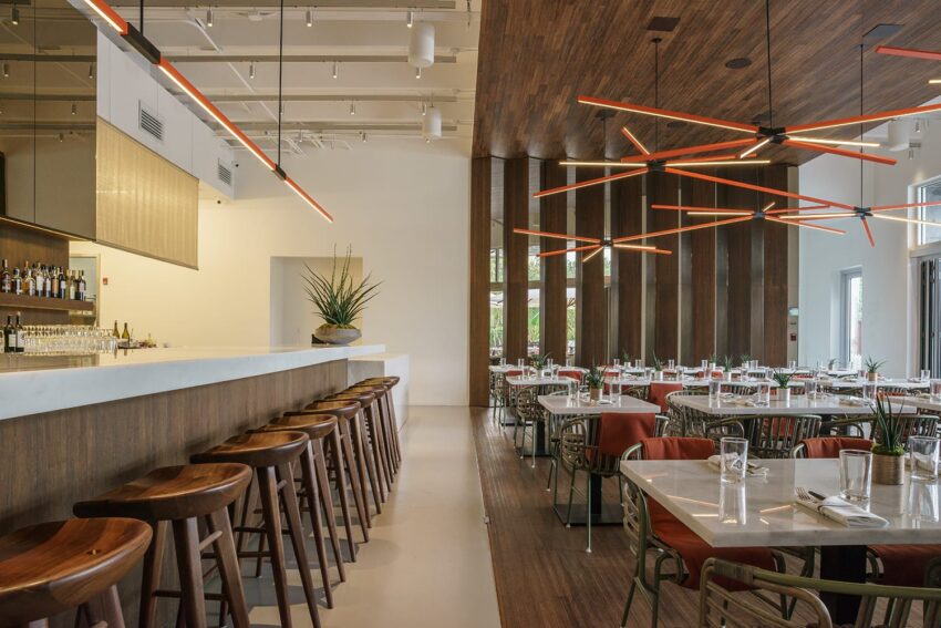 Modern restaurant interior with a sleek bar, wooden stools, and tables set for dining under artistic ceiling lights.