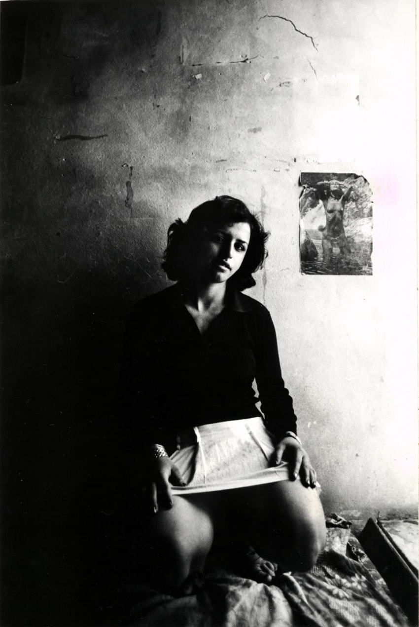 Woman sitting on a bed in a dimly lit room, holding a paper with a wall poster in the background.