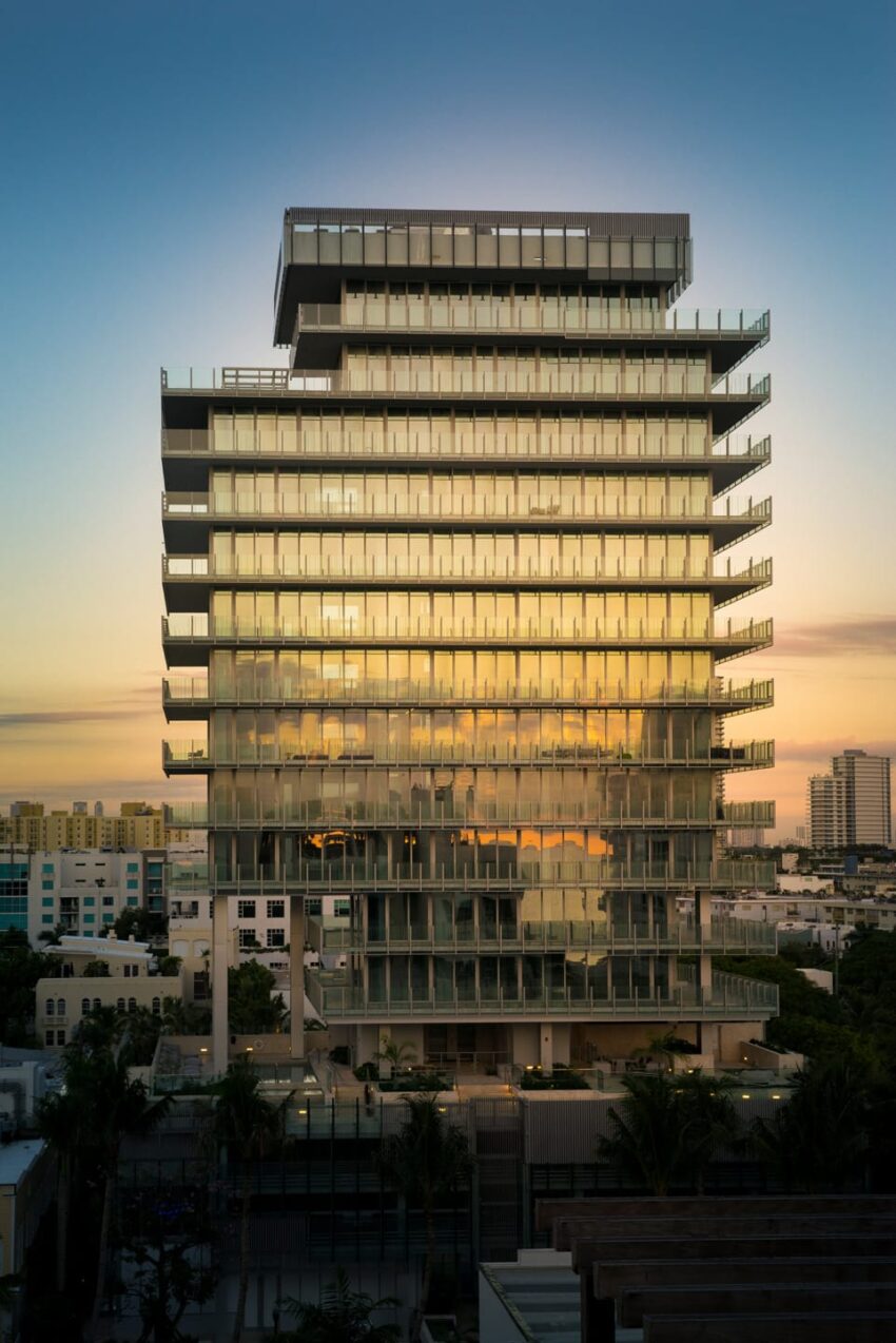 Modern glass building with multiple balconies at sunset, reflecting the sky, surrounded by urban landscape and greenery.