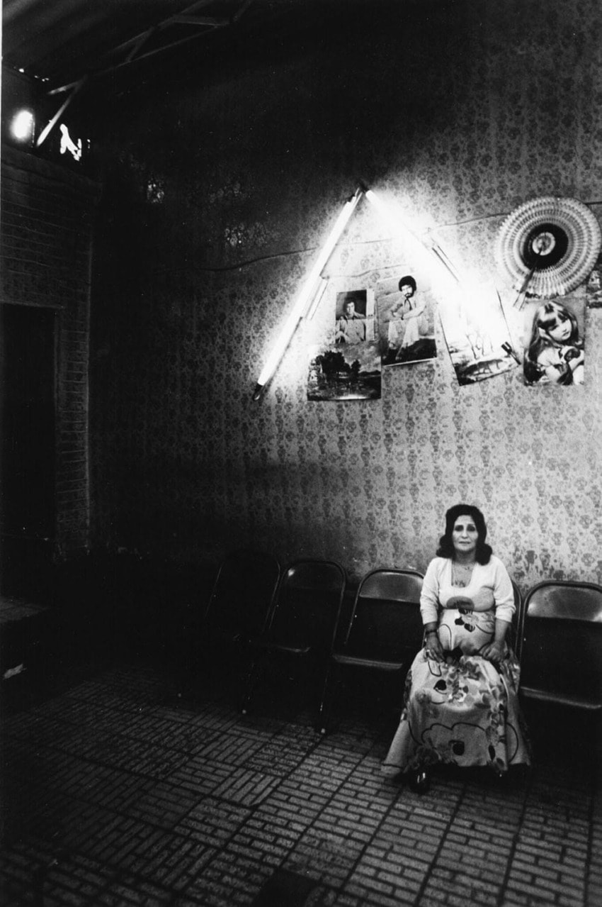 Woman sitting on a chair in a dimly lit room with vintage photos and neon lights on the wall.