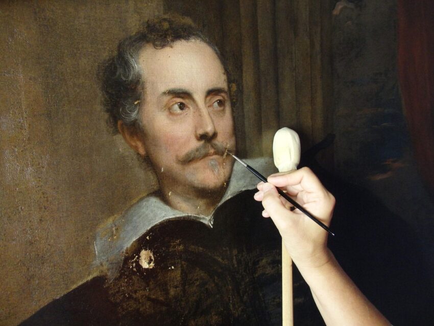 Restorer carefully working on a classical painting of a man with a mustache using a brush.