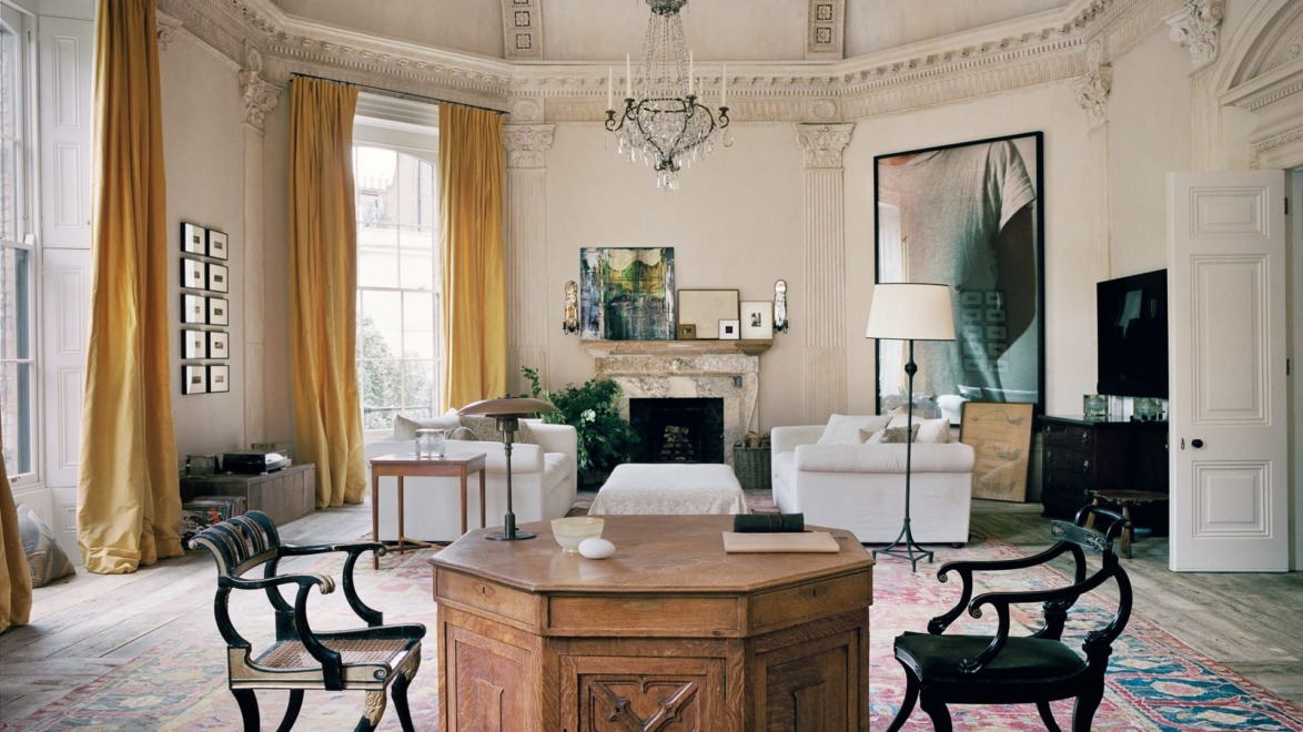 Elegant living room with ornate ceiling, chandelier, and bright yellow curtains, featuring a wooden table and white sofas.