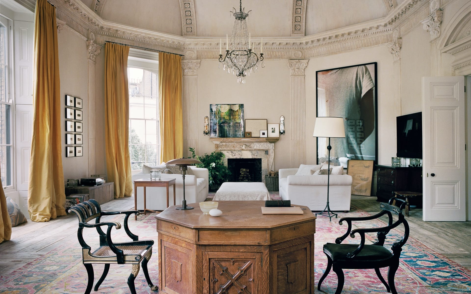 Elegant living room with ornate ceiling, chandelier, and bright yellow curtains, featuring a wooden table and white sofas.