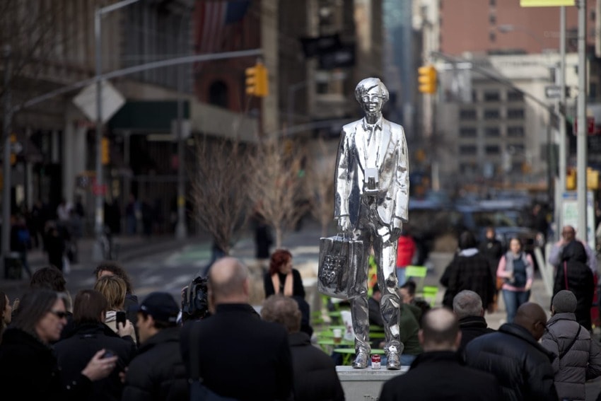 Silver statue of a man in a suit holding a bag on a busy city street with pedestrians and buildings in the background.