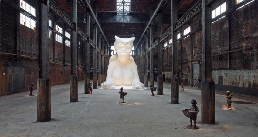 Large white sphinx-like sculpture in a dimly lit industrial space with small figures around it.