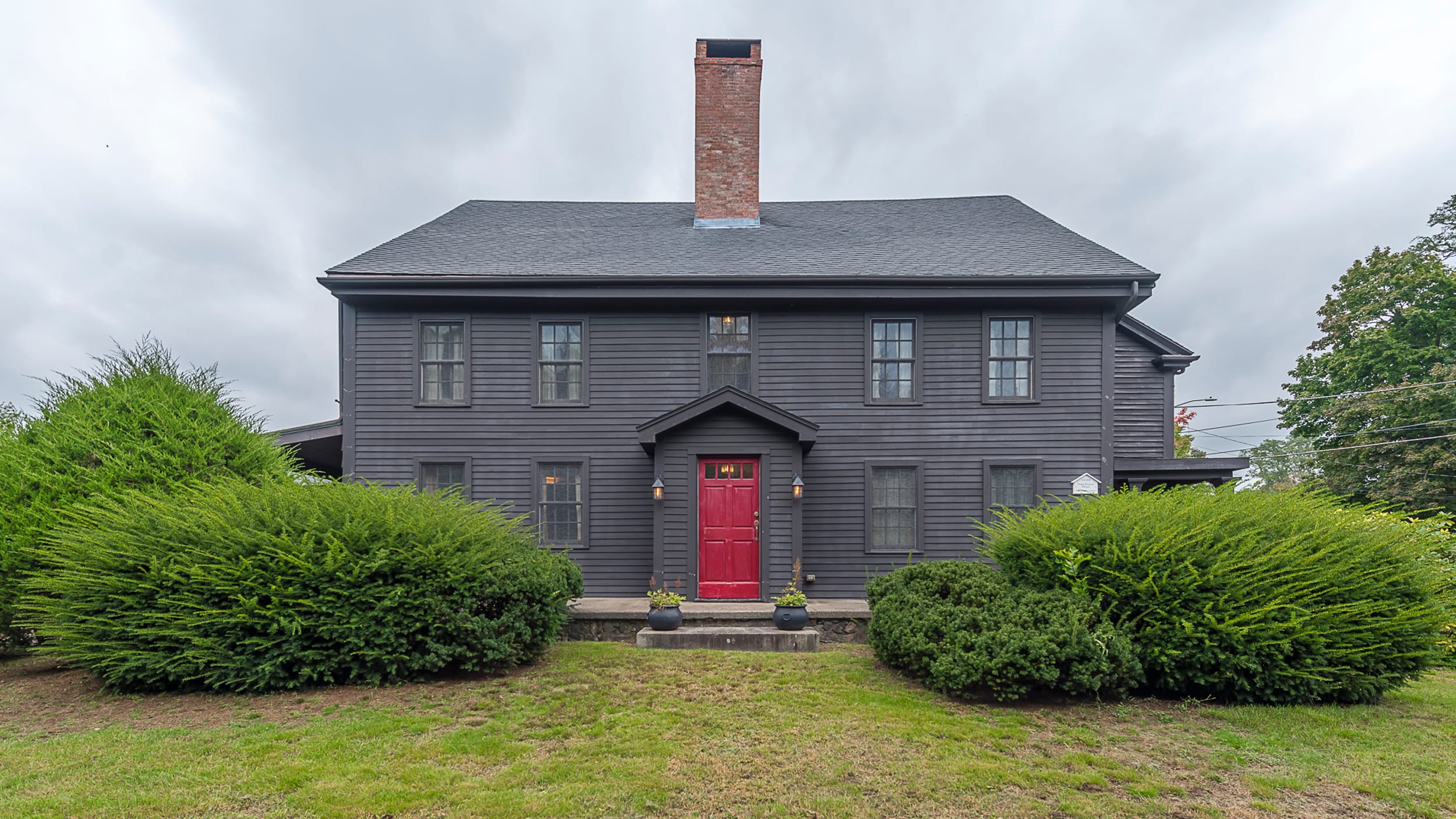 Gray two-story house with a red door, large central chimney, and hedges in front, set against a cloudy sky.