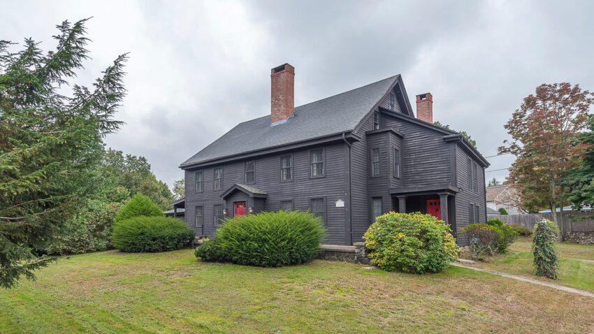 Historic large dark gray colonial house with chimneys surrounded by greenery on a cloudy day.