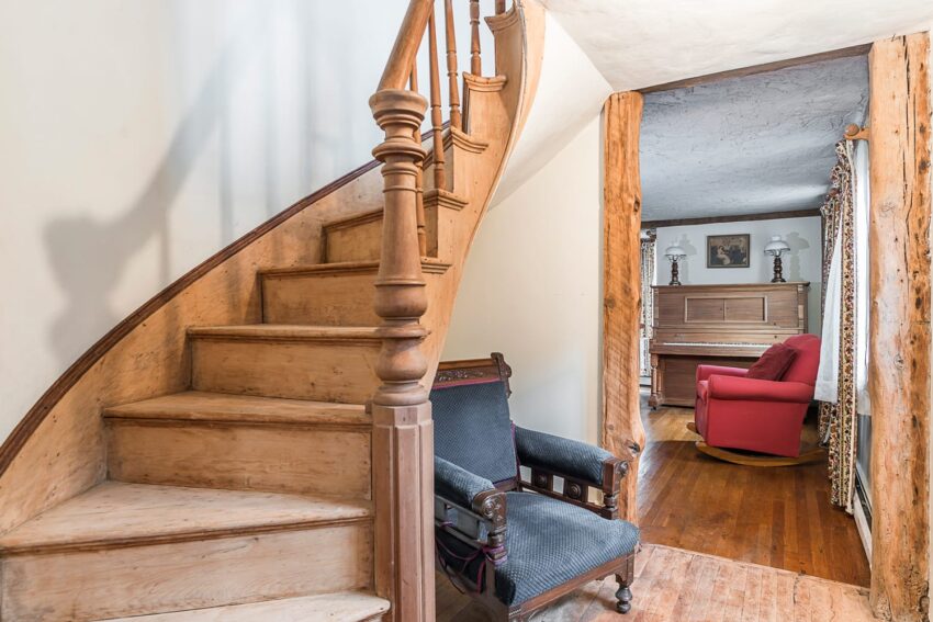 Wooden spiral staircase beside a vintage armchair leading to a cozy room with a red armchair and a piano.