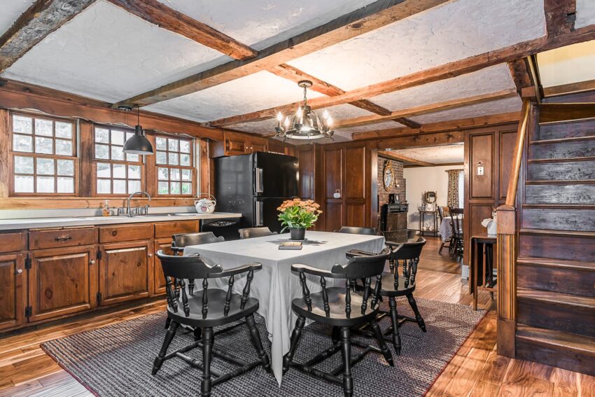 Rustic kitchen with wooden beams, dining table, black chairs, wood cabinetry, and a chandelier, creating a cozy atmosphere.
