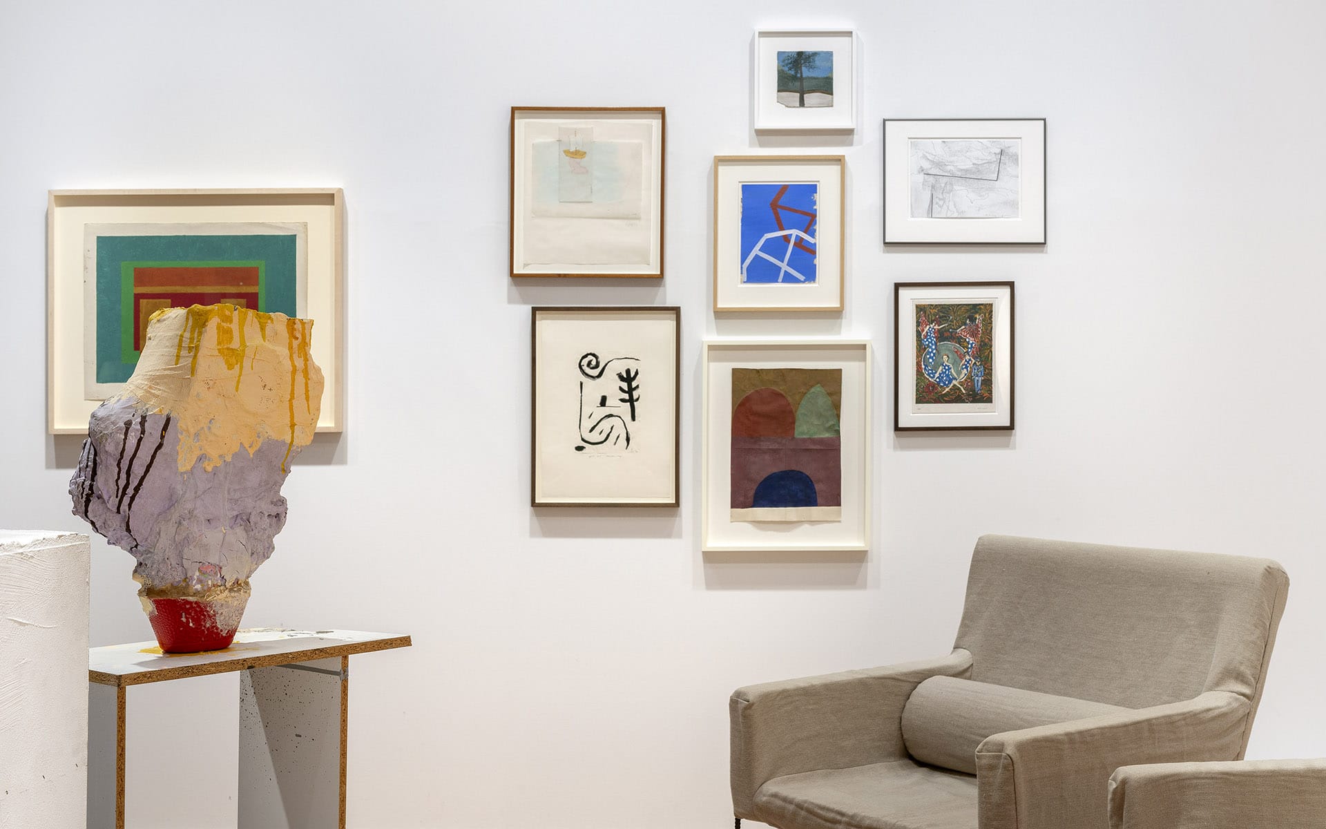 Art gallery wall with various framed abstract paintings and a clay sculpture; gray armchair in the foreground.