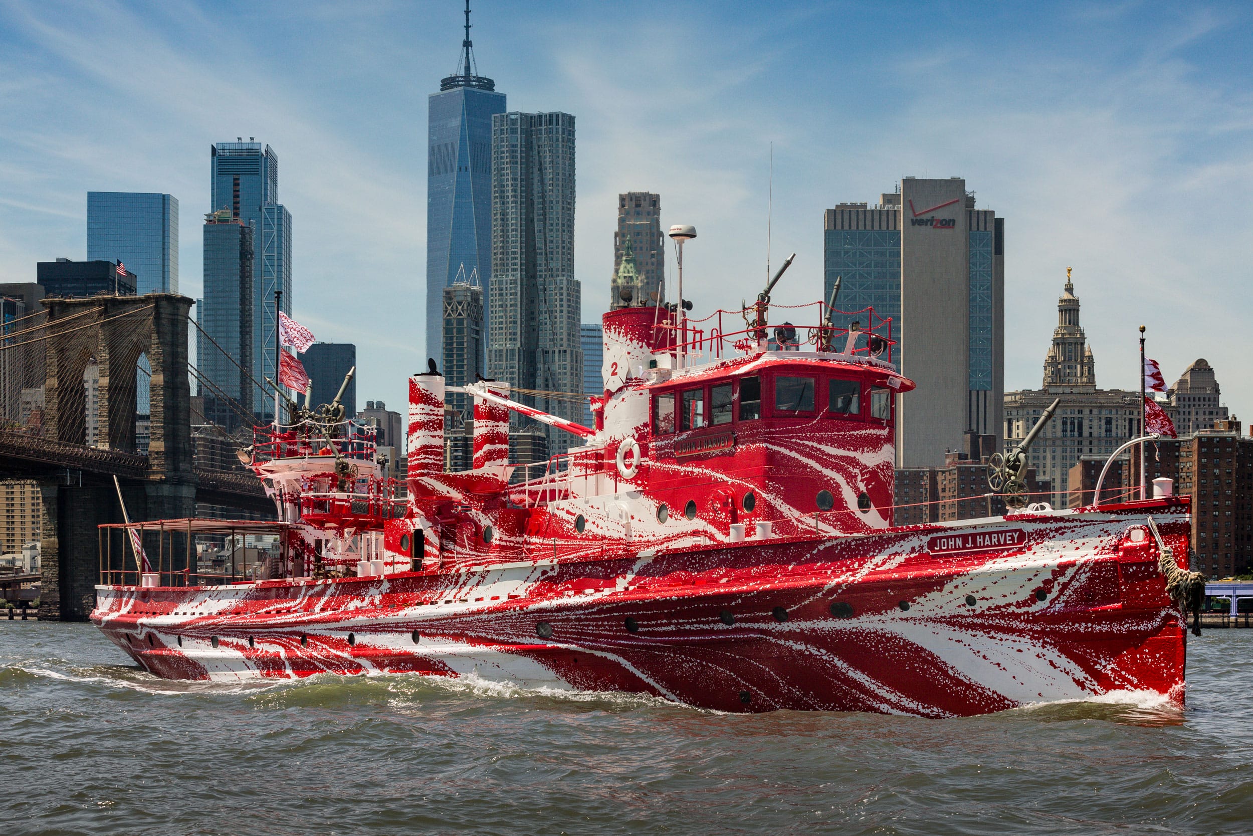 Red and white camouflaged ship sailing in a city harbor with skyscrapers and a bridge in the background.