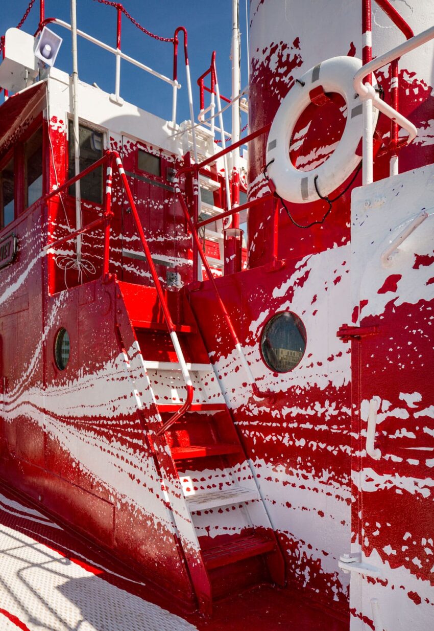 Red and white ship exterior with peeling paint and circular windows, featuring a life preserver and railing.