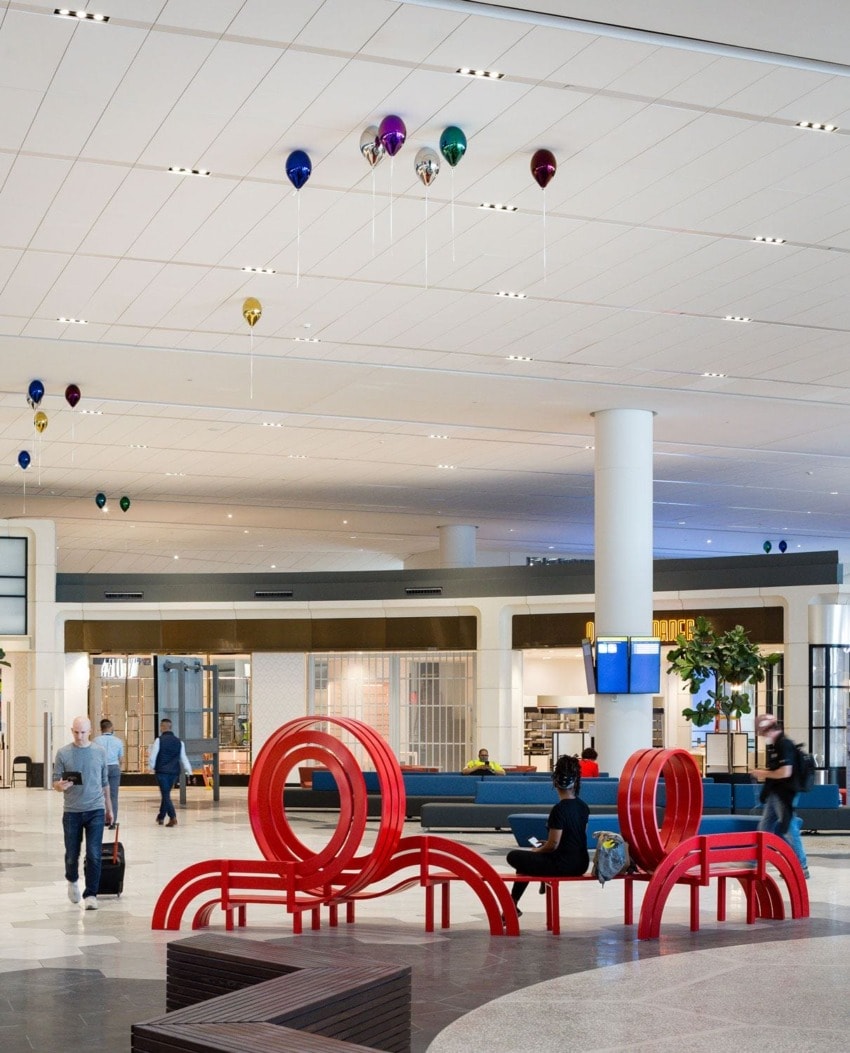 Modern airport terminal with red abstract seating and passengers moving through the space, colorful balloons hang from ceiling.