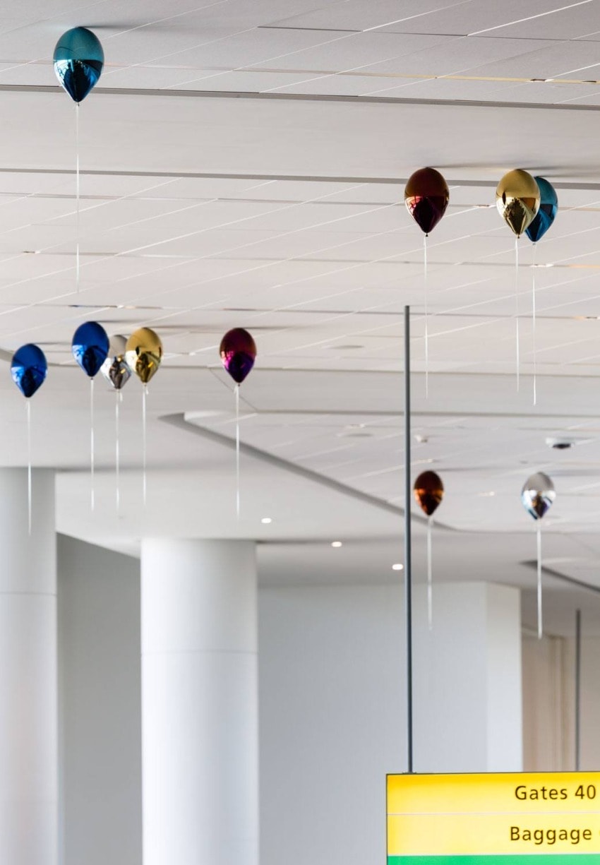 Colorful balloons floating near the ceiling in an airport terminal with a sign indicating gates and baggage in the foreground.