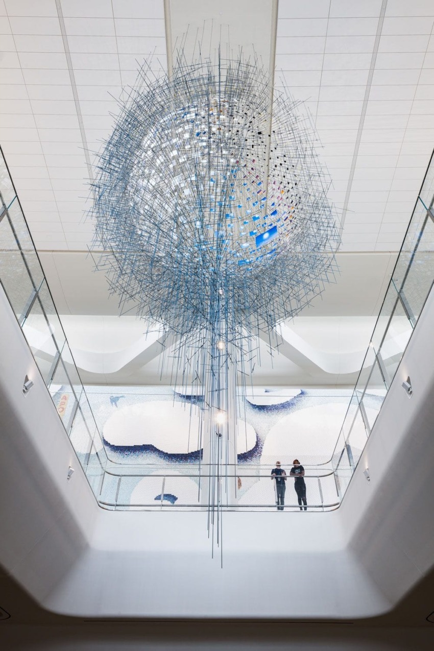 Large abstract sculpture hanging in a modern atrium with two people standing on a balcony below, observing the artwork.