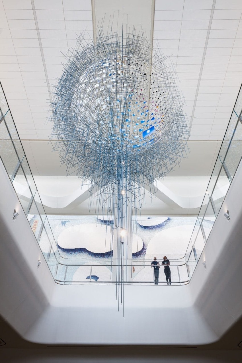 Large abstract sculpture hanging in a modern atrium with two people standing on a balcony below, observing the artwork.