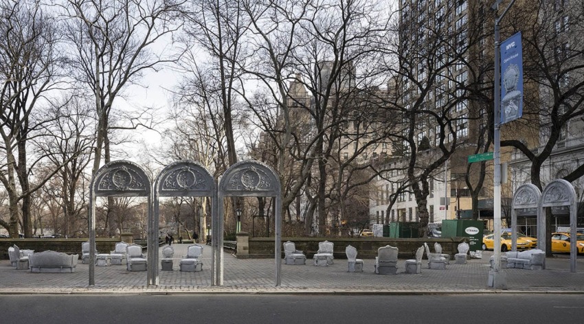 Outdoor art installation featuring metallic arches and sculptures on a city street, with bare trees and taxis in the background.