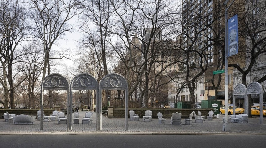 Outdoor art installation featuring metallic arches and sculptures on a city street, with bare trees and taxis in the background.