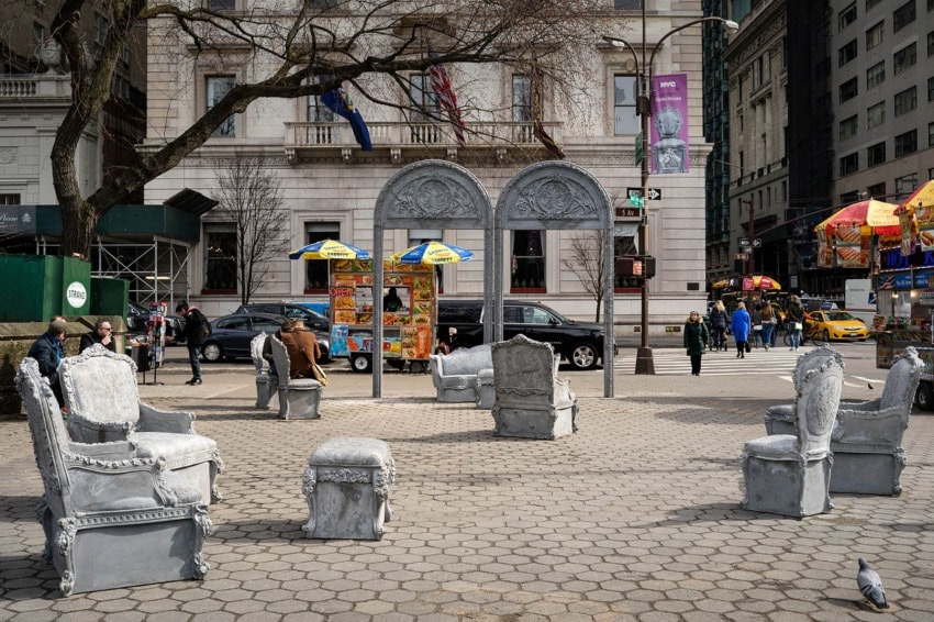 Public art installation featuring ornate stone chairs and arches in a park setting with people and city buildings in the background.