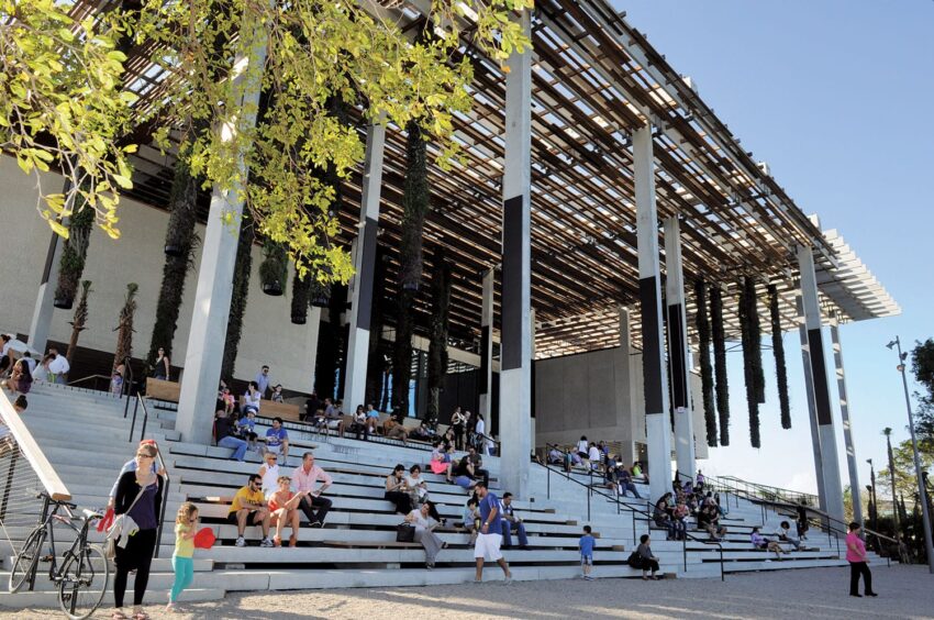 People sitting on steps outside a modern building with tall columns and hanging plants, under a clear blue sky.