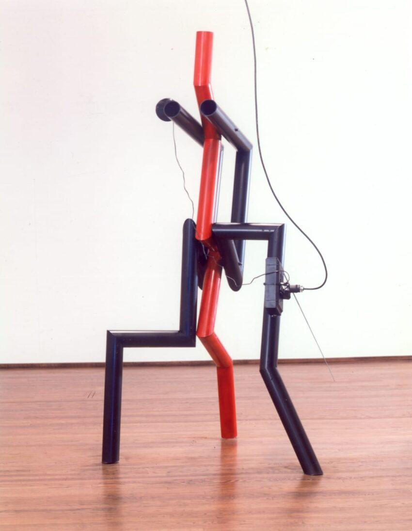 Abstract metal sculpture with red and black interconnected tubes on a wooden floor.